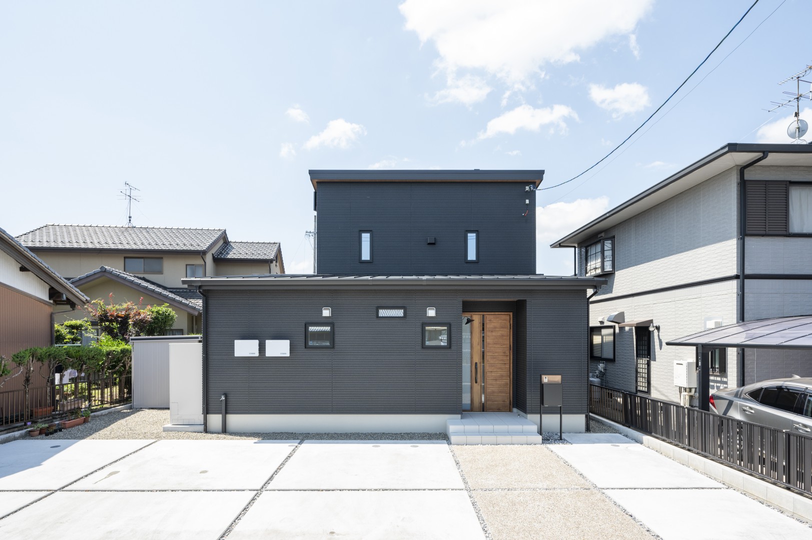 濃いチャコールの外壁の2階建て。道路側の窓は小さなものが数か所だけで、中央に木目の玄関ドア。手前に駐車スペースと境界沿いのフェンスがある
