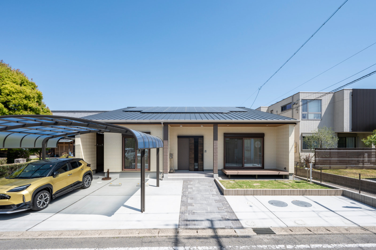ベージュの外壁の平屋。金属屋根に太陽光パネルが載り、左にカーポート、右にウッドデッキと芝生。両隣に建物が接している